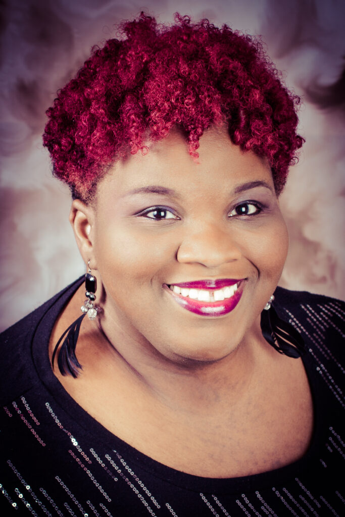 Image of Angel Austin, an infinifat Black person with short, bright pink, tight curls, smiling brightly at the camera.